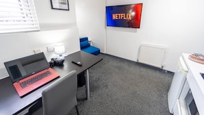 Una habitación con una computadora portátil, un control remoto, una cámara, una silla azul, un televisor con 'NETFLIX' y un radiador.