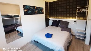 A compact hotel room with a bunk bed, a large bed with a white duvet, a wall-mounted shelf, and a mirror.