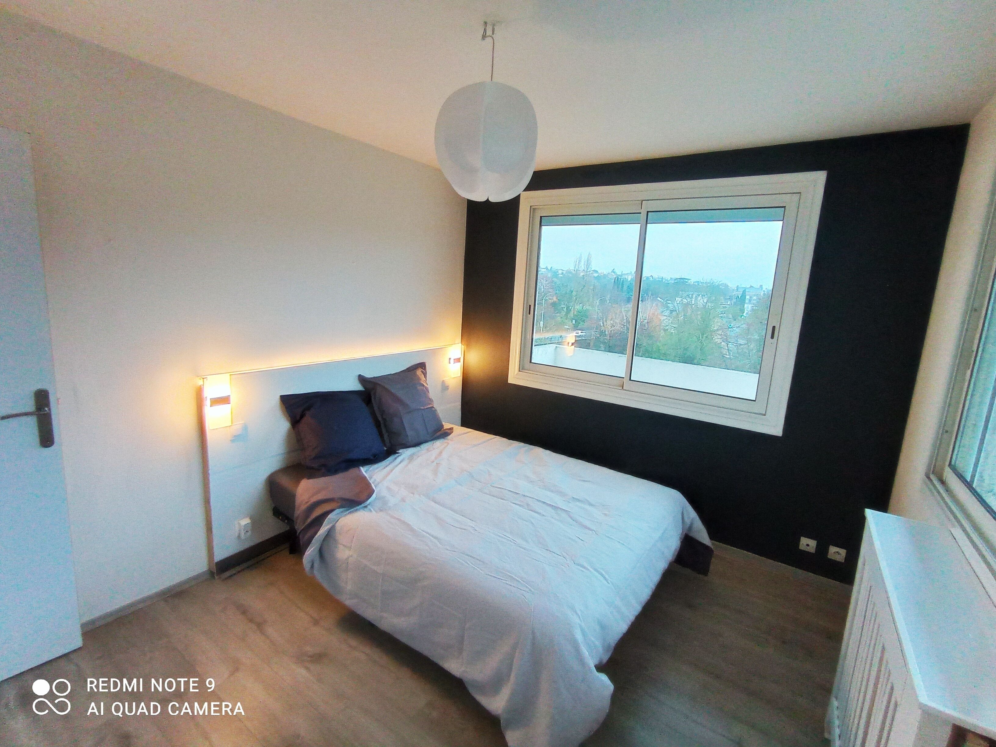 Ein modernes Schlafzimmer mit einem großen Bett, einem Fenster mit Blick auf die Stadt und einer Deckenleuchte.