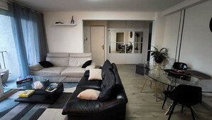 A modern living room with a black leather sofa, a glass coffee table, and a beige sofa. There is a dining area with a glass table and chairs. The room has a large window with curtains.