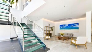 Un intérieur moderne avec un escalier en verre, un espace de vie comprenant un canapé et une table basse, ainsi qu’un grand tableau représentant une scène de plage.