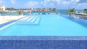 Une piscine d’un bleu éclatant, bordée de carreaux, entourée de chaises longues et de palmiers, avec une vue sur la mer et les bâtiments en arrière-plan.