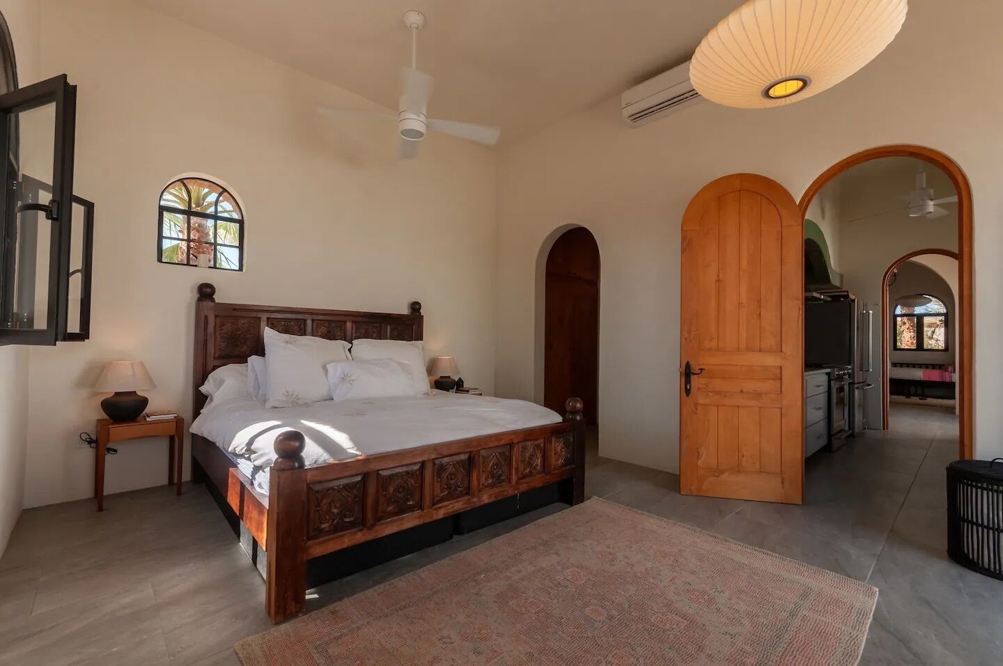 Une chambre à coucher avec un lit en bois, une table de chevet, une lampe, un ventilateur de plafond, une fenêtre et une porte donnant sur la cuisine.