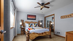 Une chambre à coucher avec un lit en bois, un ventilateur de plafond, une grande fenêtre avec des rideaux et un tableau accroché au mur.
