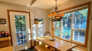 Une cuisine avec une grande table en bois, deux tabourets de bar, un luminaire suspendu et une vue sur l’extérieur grâce à de grandes fenêtres.