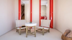 Une petite pièce avec deux chaises en osier et une table ronde, une porte à cadre rouge et un panier en osier posé sur le sol.