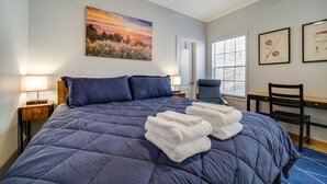 Una cama bien hecha con un edredón azul, dos almohadas y toallas blancas dobladas. En la pared, un cuadro de un atardecer, una silla y un escritorio con una silla.