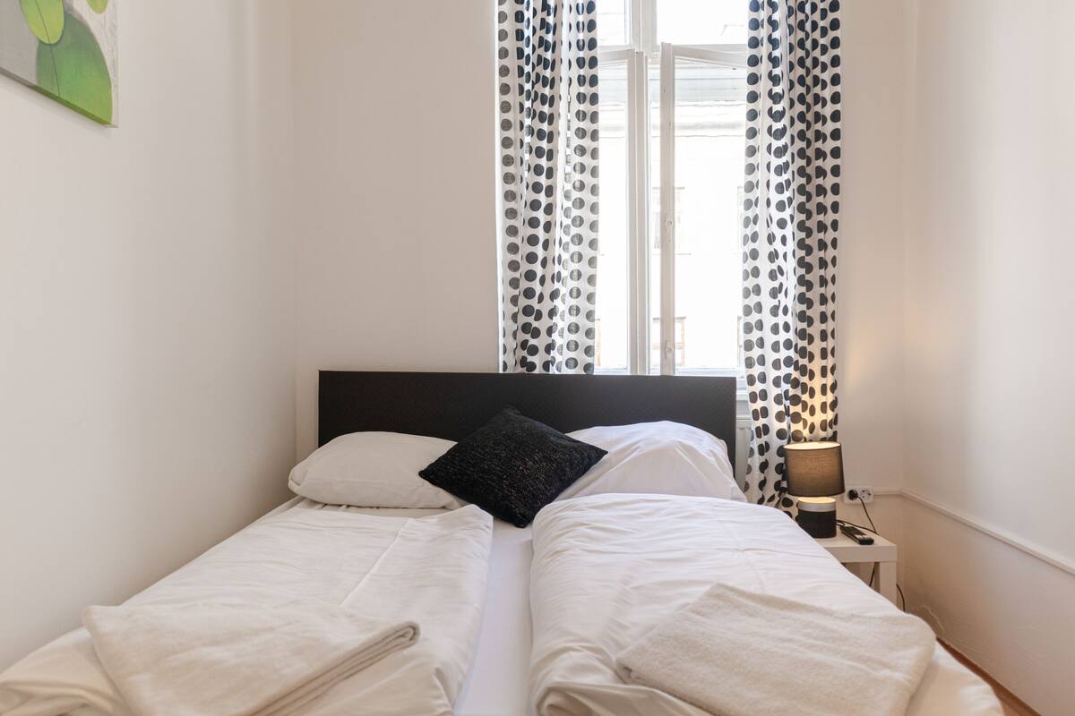 A neatly made twin bed with white linens, a black pillow, and a small bedside table with a lamp.