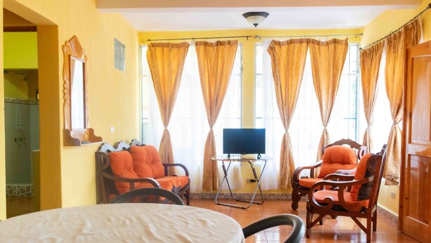Una habitación con una mesa redonda, dos sillas de madera, un televisor sobre un mueble bajo y cortinas en las ventanas.