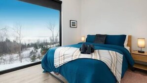 Une chambre avec une grande fenêtre, un lit recouvert d’une couverture bleue, une table de chevet avec une lampe et un tableau encadré accroché au mur.
