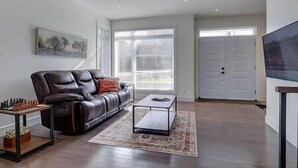 A living room with a leather sofa, a coffee table, a TV on a stand, and a door with a window. - Hôtel à la maison - Le Faucon Azuré, golf et ski (Potton)