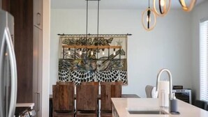A modern kitchen with wooden chairs, a central island, pendant lights, and a decorative wall piece. - Hôtel à la maison - Le Faucon Azuré, golf et ski (Potton)