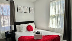 A neatly arranged bedroom with a bed, a bedside table, a nightstand, a red throw blanket, and a small basket with toiletries. - 💎 Chic Private Room 🛜 Fast Wi-Fi 👣Near LIRR & Shops (Hicksville)