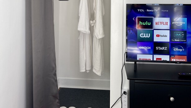 A bathroom with a shower curtain, two white towels, and a black cabinet with a TV displaying various streaming services. - 💎 Chic Private Room 🛜 Fast Wi-Fi 👣Near LIRR & Shops (Hicksville)