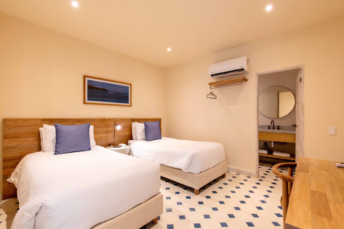 Habitación de hotel con dos camas, cabecera de madera, mesita de noche, escritorio, silla, baño con espejo y un cuadro de un barco en la pared.
