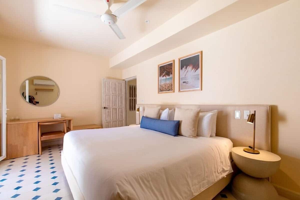 Habitación de hotel con cama, escritorio, espejo redondo, ventilador de techo y dos cuadros en la pared.