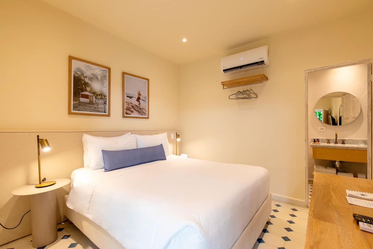 Habitación de hotel con cama, mesita de noche, lámpara, unidad de aire acondicionado y baño con espejo y lavamanos.