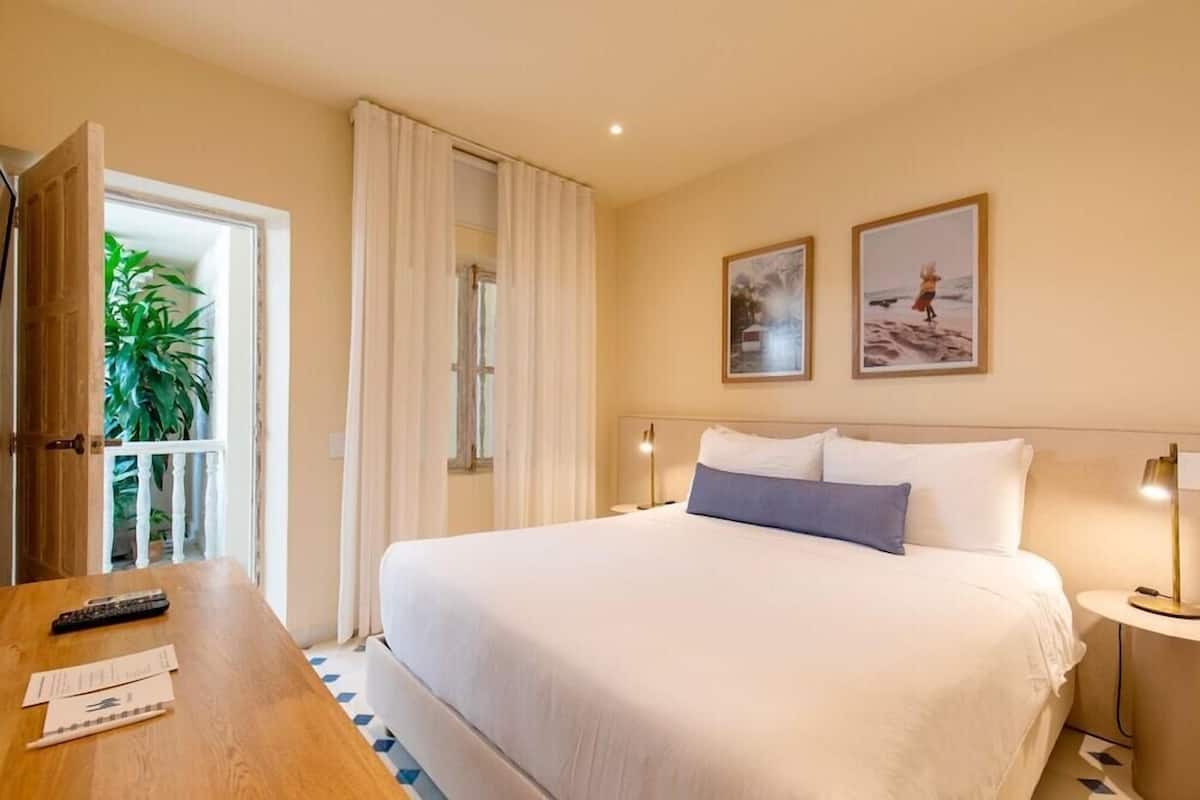 Habitación de hotel con cama, escritorio, silla, mesita de noche, lámpara y dos cuadros en la pared.