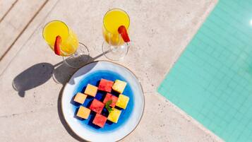 Une table au bord de la piscine avec deux verres contenant une boisson jaune, une assiette de cubes de pastèque et un verre d’eau.