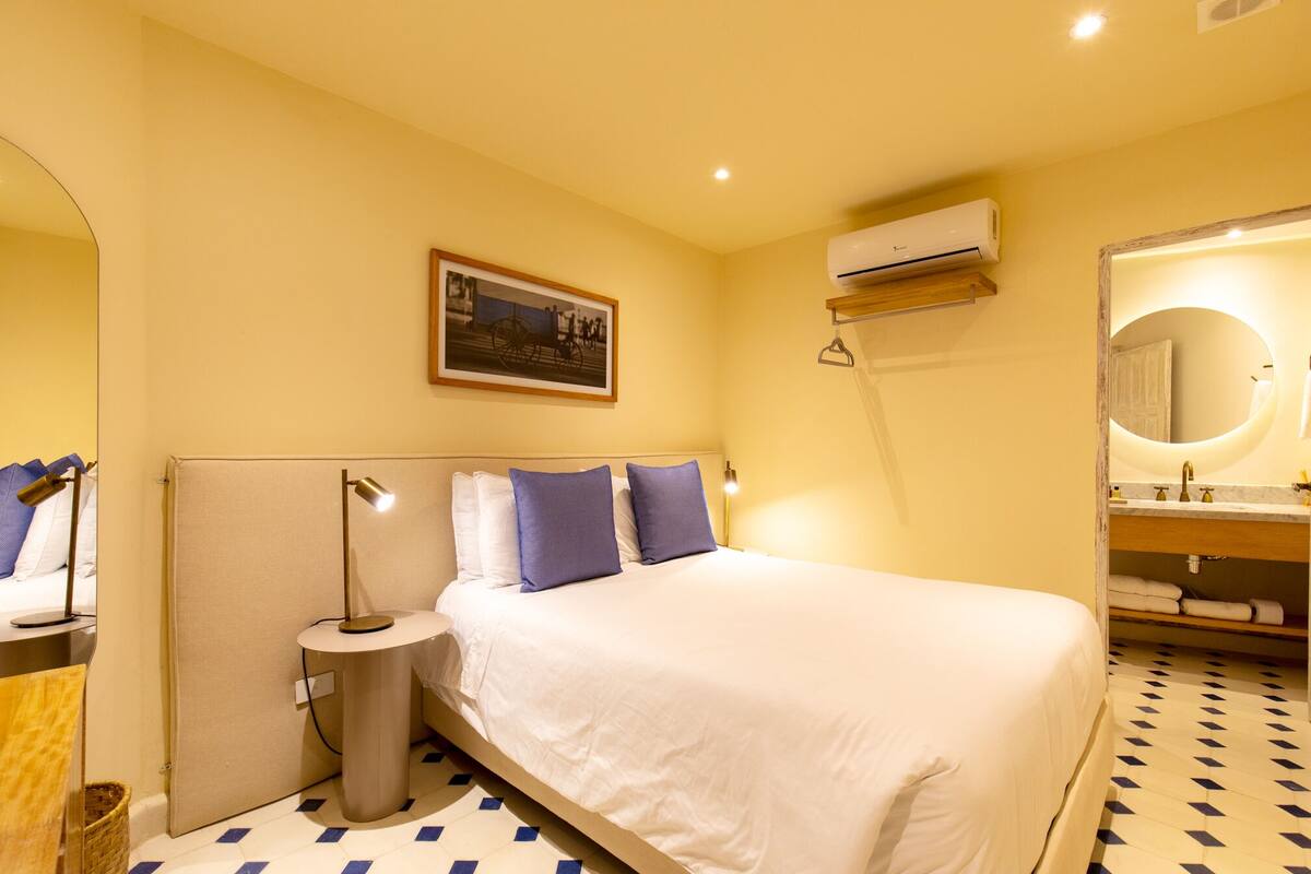 Habitación de hotel con cama, lámpara de noche, aire acondicionado y baño con espejo y lavamanos.