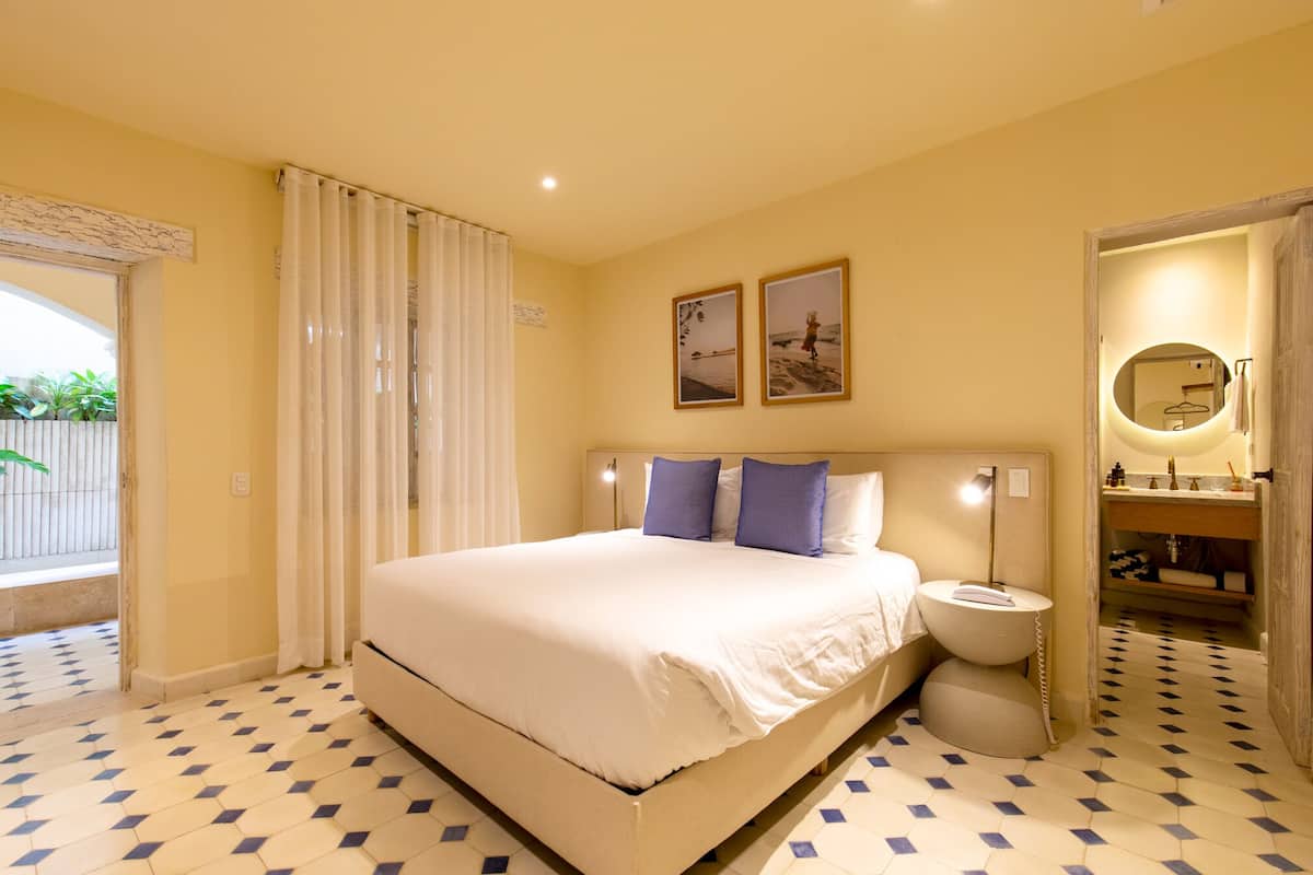Habitación de hotel con una cama grande, una mesita de noche, un baño con espejo y vistas al exterior.