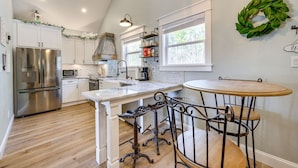 Une cuisine avec des meubles blancs, un réfrigérateur en acier inoxydable, une table à manger en bois avec des chaises en métal et une guirlande décorative accrochée au mur.
