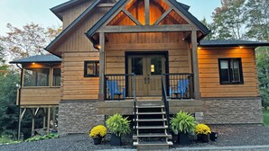 A log cabin with a stone foundation, a wooden porch with blue chairs, and a staircase leading to the entrance. - New 5 bed 4 bath cottage on Turtle Lake, Privacy! (Seguin)
