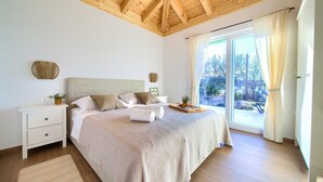 Ein Schlafzimmer mit Holzboden, einem großen Bett mit beige farbenem Bettbezug, zwei Nachttischen, einer kleinen Topfpflanze und Blick auf einen Garten durch eine Schiebetür.
