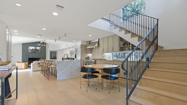 A modern kitchen with a central island, a dining area with chairs, and a staircase with a metal railing.