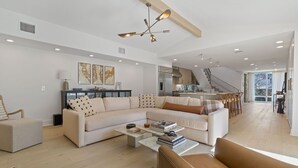 A spacious living area with a large L-shaped sofa, a coffee table, and a brown leather armchair. A kitchen is visible in the background with a staircase leading to another level.