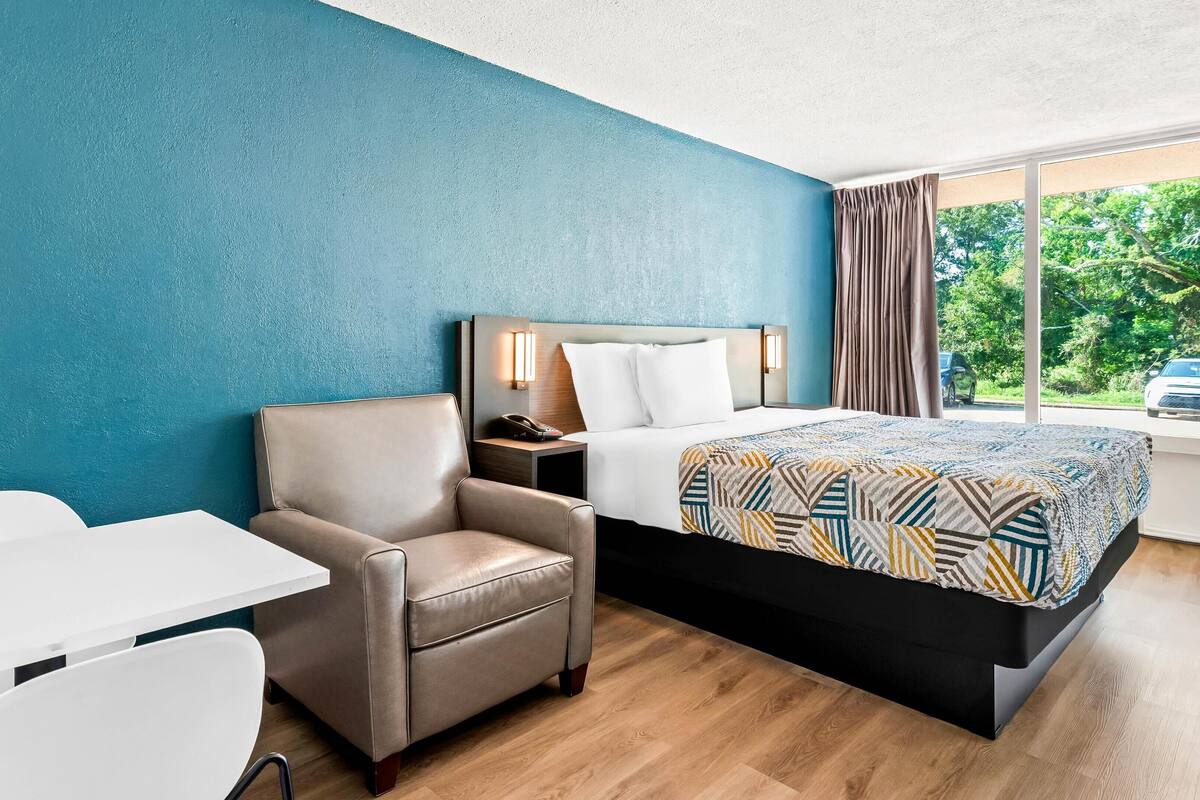 A hotel room with a bed, a chair, a desk, and a window with a view of greenery and a car.