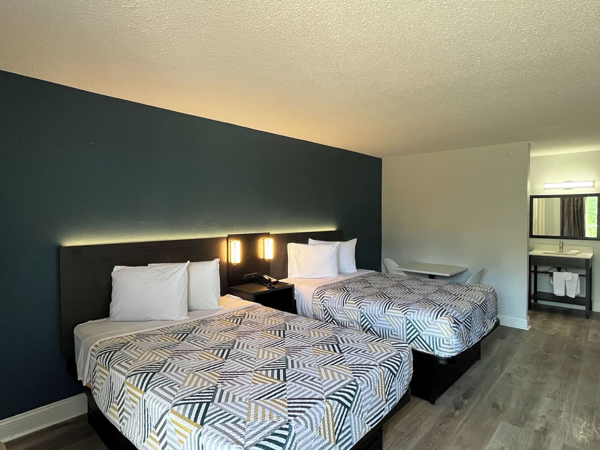 A hotel room with two beds, a bedside table with a lamp, a small desk, and a bathroom with a sink and mirror.