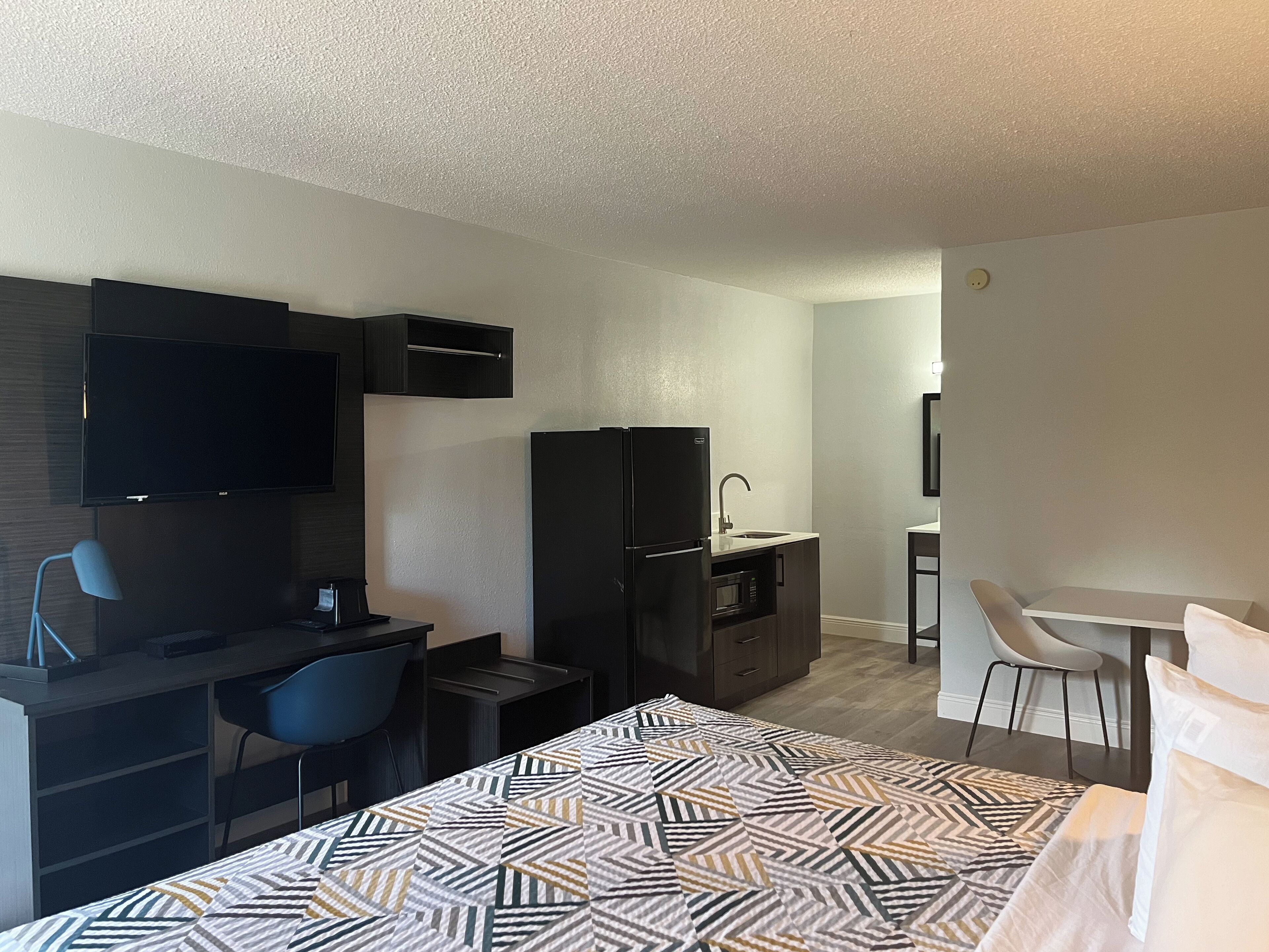 A hotel room with a bed, a desk with a chair, a TV, a refrigerator, a microwave, and a small kitchenette.