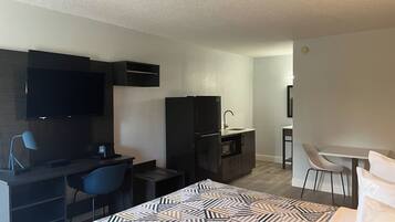 A hotel room with a bed, a desk with a chair, a TV, a refrigerator, a microwave, and a small kitchenette.