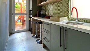 Cozinha com bancada branca, pia e janela. Há dois bancos e uma porta que dá para uma área externa.