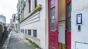 A narrow street with buildings on either side. There are cars parked along the street, and a red door is visible on the right side. - Nomad's Family - 3BDR Charming Hideout Porto (Porto)