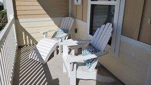 A covered outdoor seating area with two Adirondack chairs and a small table, against a beige wall with a window and a decorative anchor.
