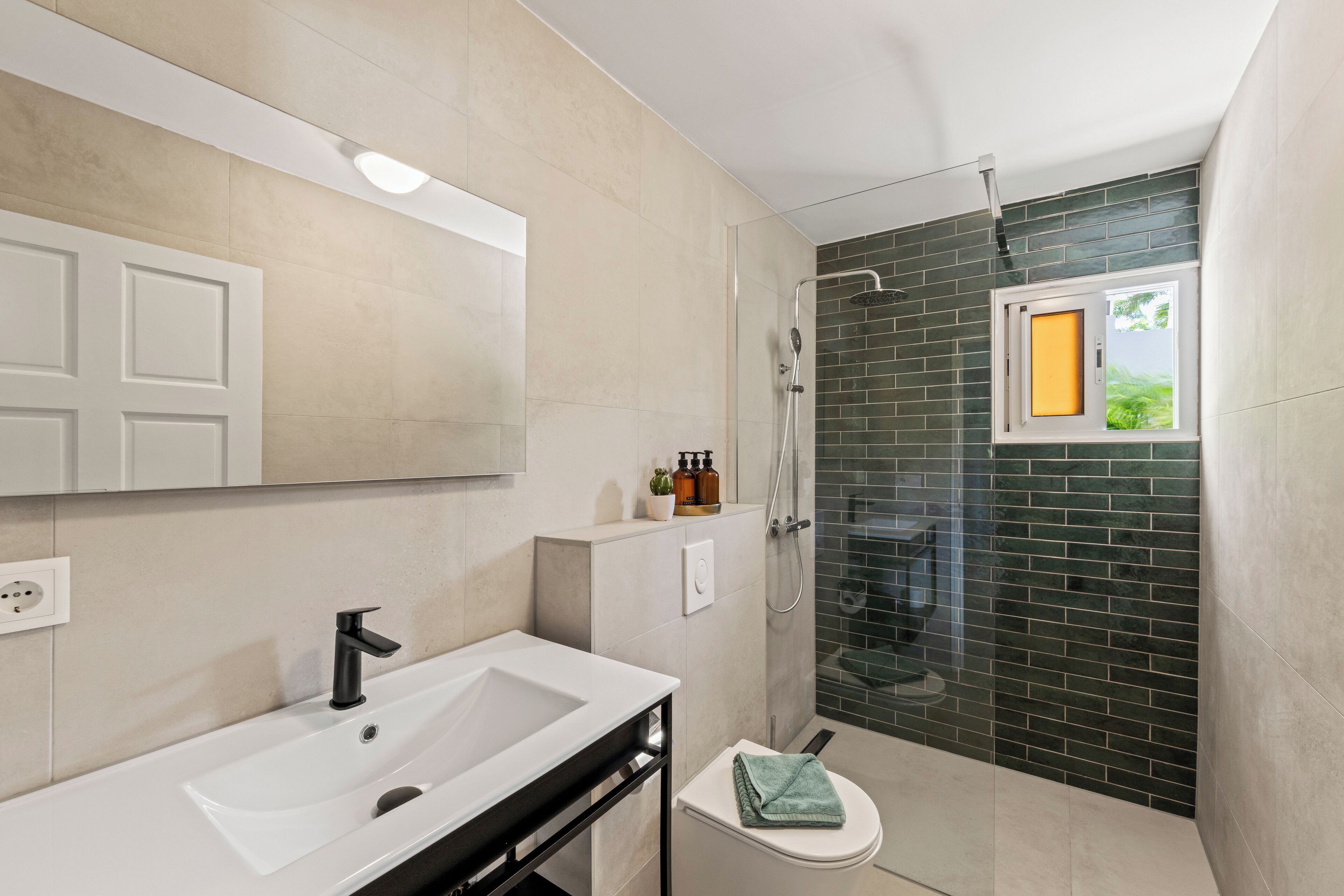 A modern bathroom with a white sink, a glass shower enclosure, and a small window with a view of greenery.