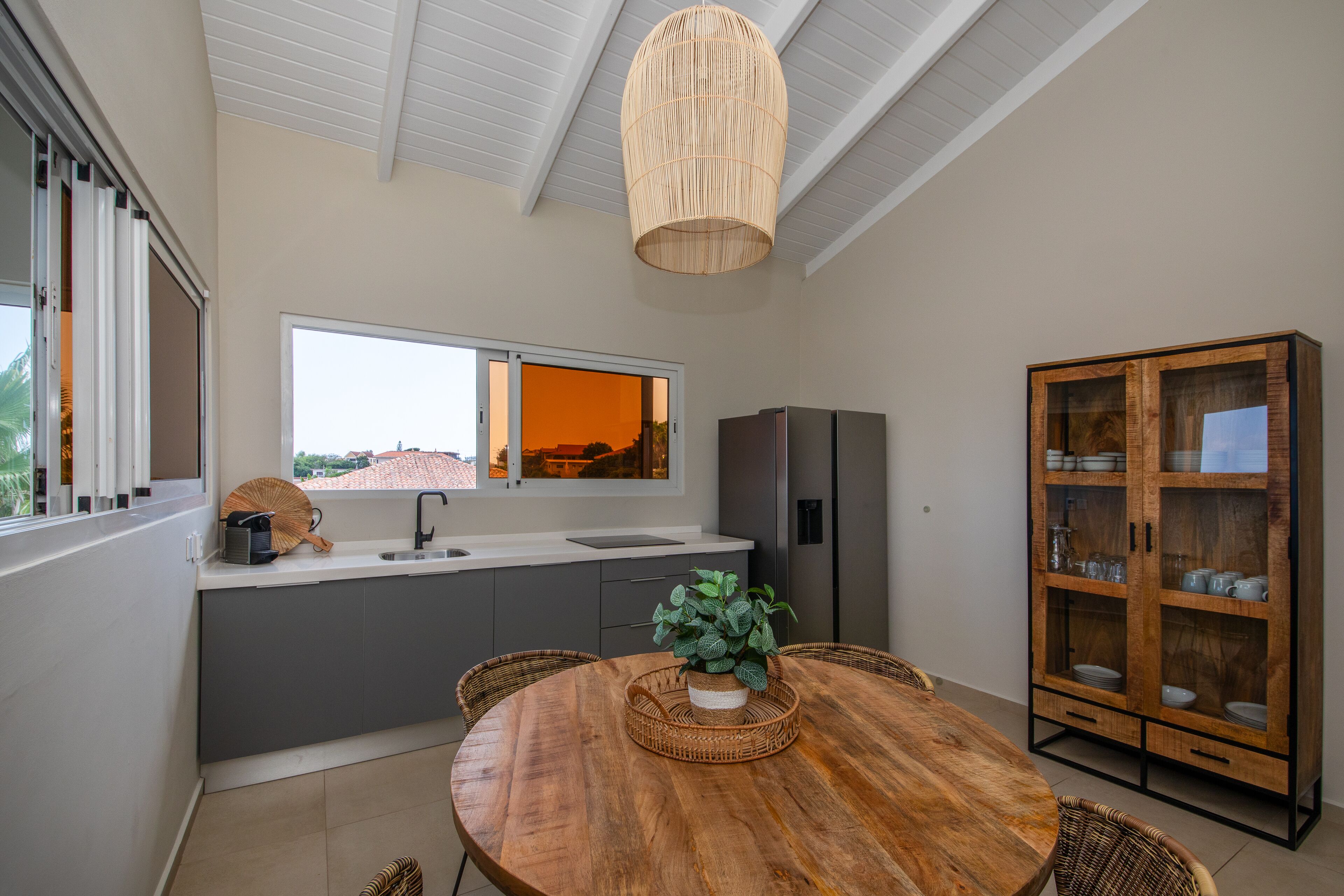 Een moderne keuken met een eethoek, met een groot raam, een hanglamp en een houten kast met glazen deuren.