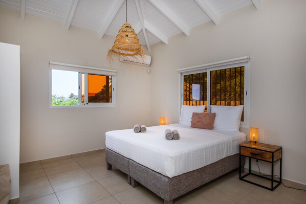 A bedroom with a large bed, two windows, a hanging light fixture, and a bedside table with a lamp. - JT Apartments Jan Thiel  (Jan Thiel)