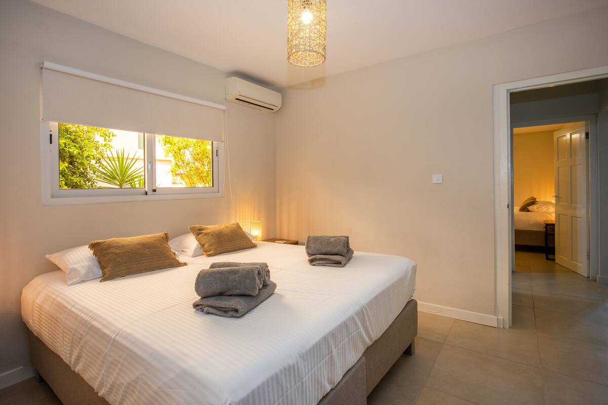 Een slaapkamer met een groot bed, een raam met uitzicht op groen, een airconditioning en een deur naar een andere kamer.