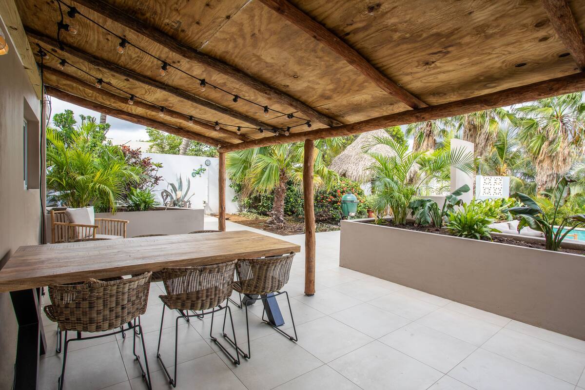 Een terras buiten met een houten tafel en rieten stoelen, omgeven door weelderig groen en een constructie met rieten dak.