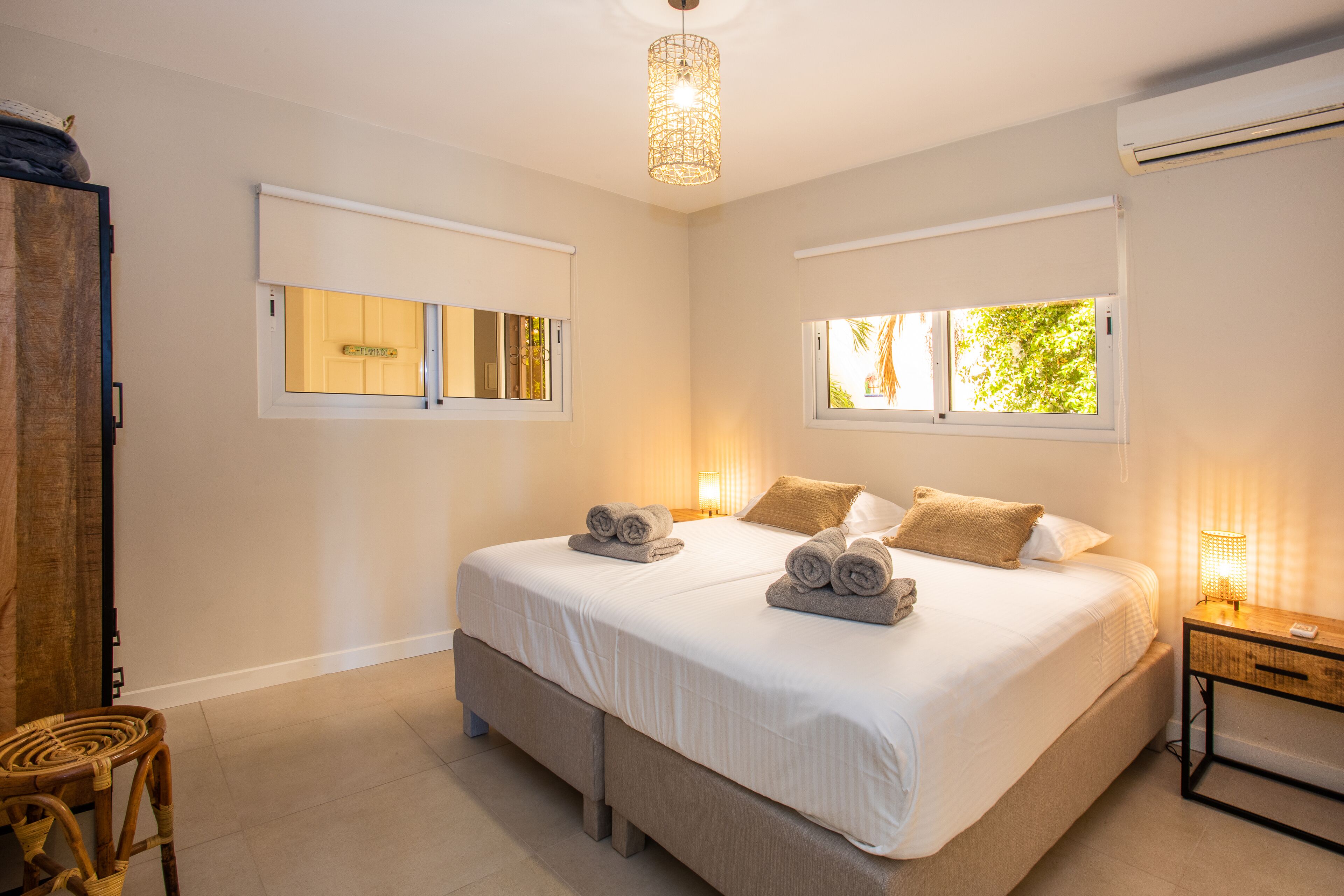 A bedroom with a large bed, two bedside tables, a hanging light fixture, and a window with a view of trees.