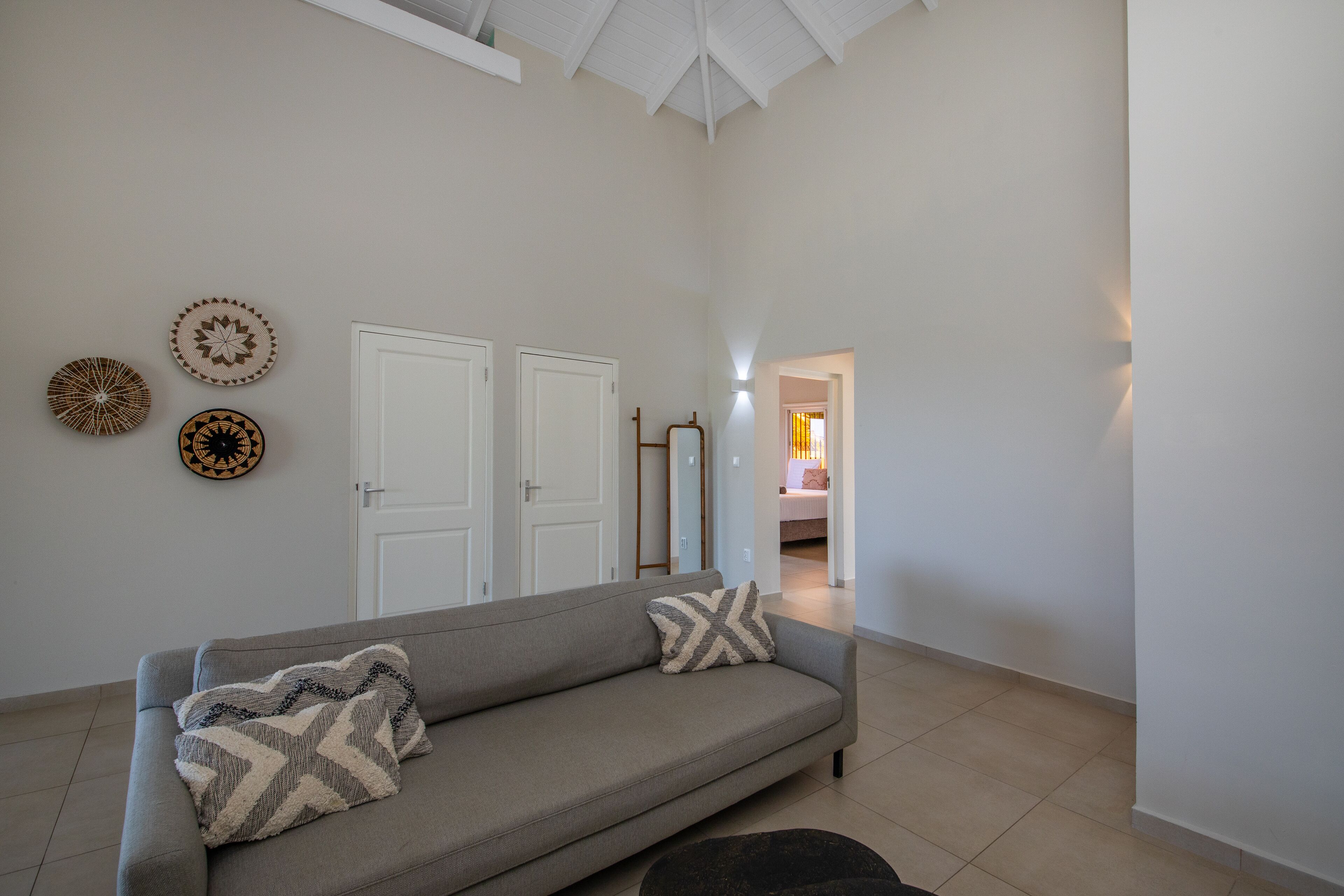 A living room with a grey sofa, two patterned throw pillows, and three wall decorations. There are two white doors and a hallway leading to another room.