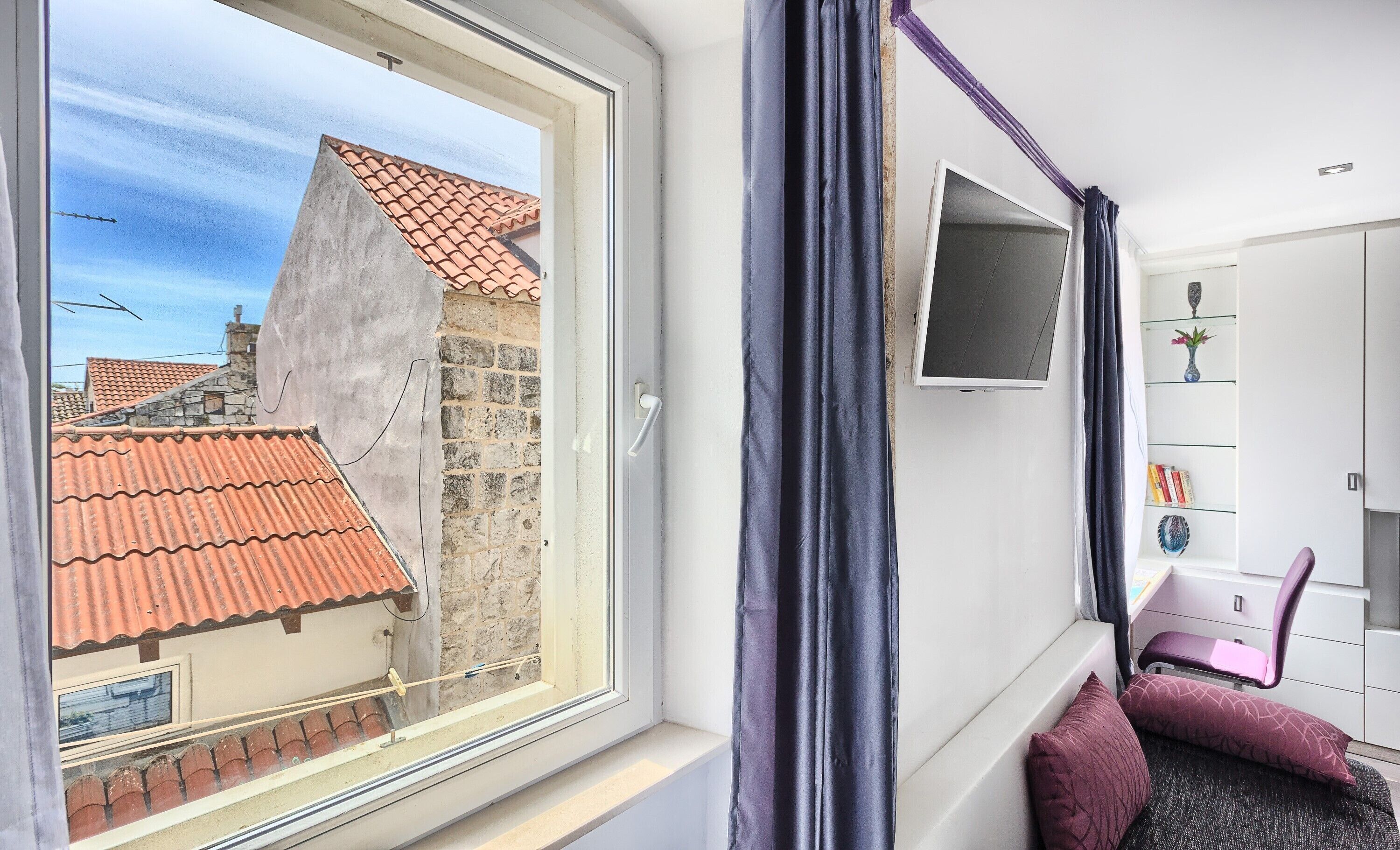 A room with a large window, a flat-screen TV mounted on the wall, a pink chair, and a small shelf with decorative items.