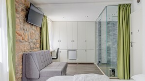 A modern hotel room with a stone accent wall, a grey sofa, a bed with patterned bedding, a flat-screen TV, and a glass-enclosed shower area.