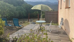 A wooden deck with a table and chairs, a green umbrella, and a view of hills in the background. - Studio Figari, au Calme Avec Magnifique vue sur la Montagne et les Vignes (Figari)