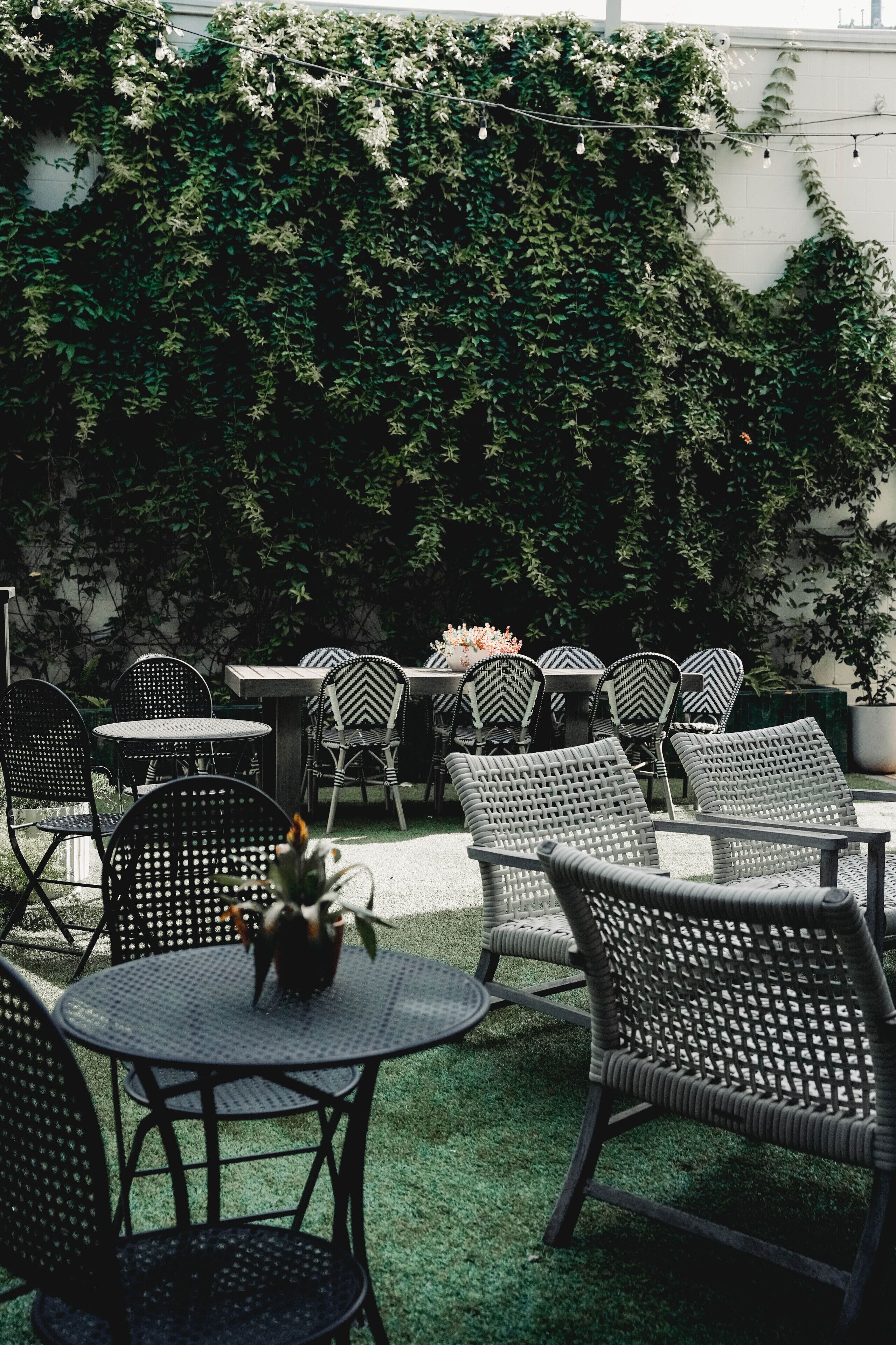 An outdoor patio with wicker furniture, tables, and chairs, surrounded by lush greenery and a white wall.