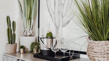 Una encimera con un balde de metal, dos vasos y diversas plantas, incluyendo un cactus grande y una planta verde alta.