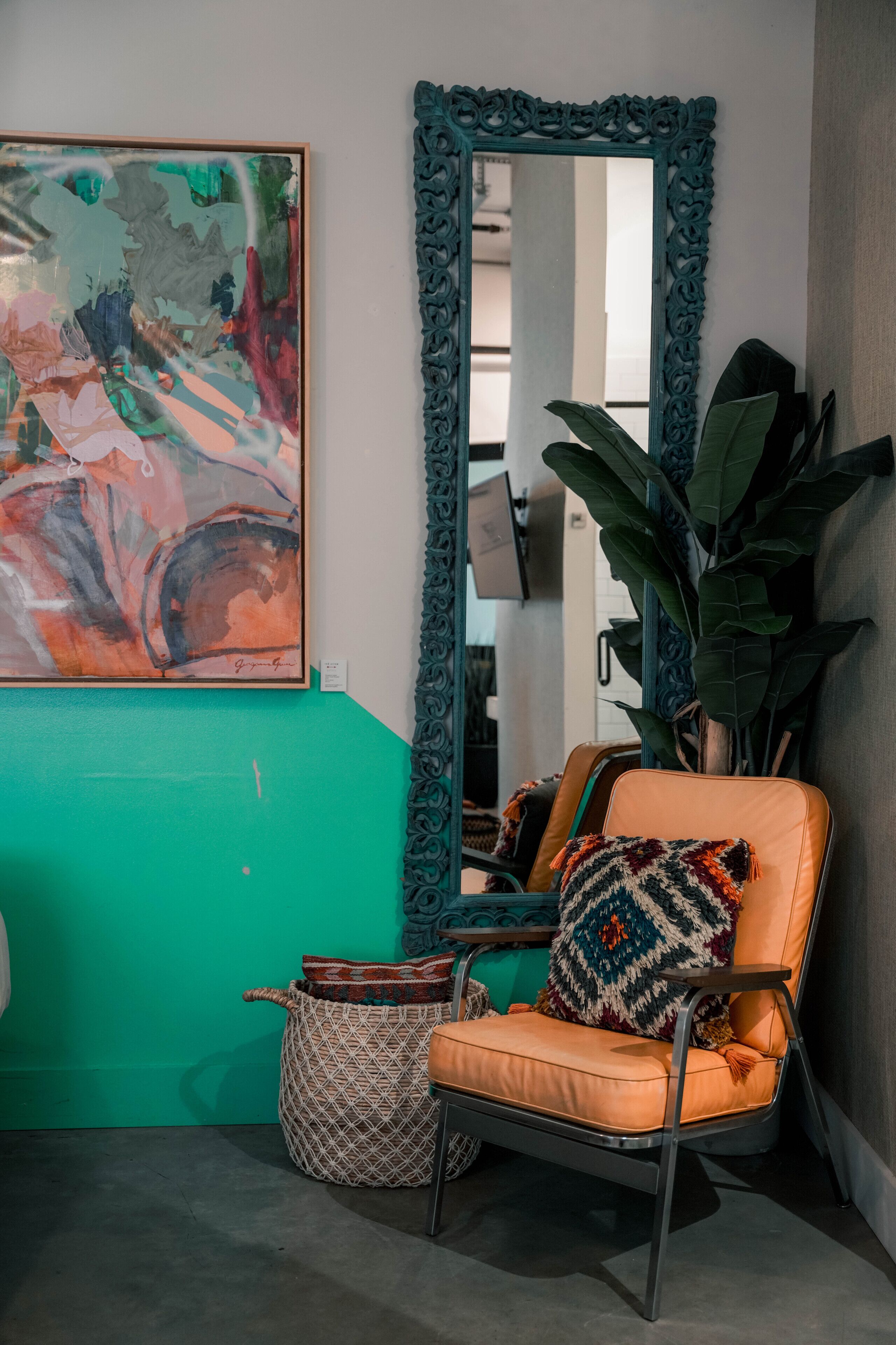 A room with a teal accent wall, a large abstract painting, a mirror with an ornate frame, a plant, and a chair with a patterned cushion.